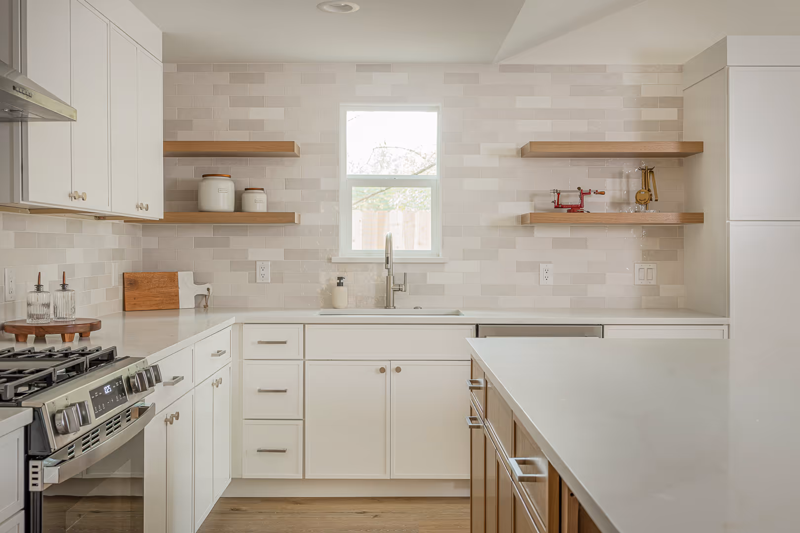 Modern kitchen with white cabinetry, wooden floating shelves, stainless steel stove, and a window above the sink.