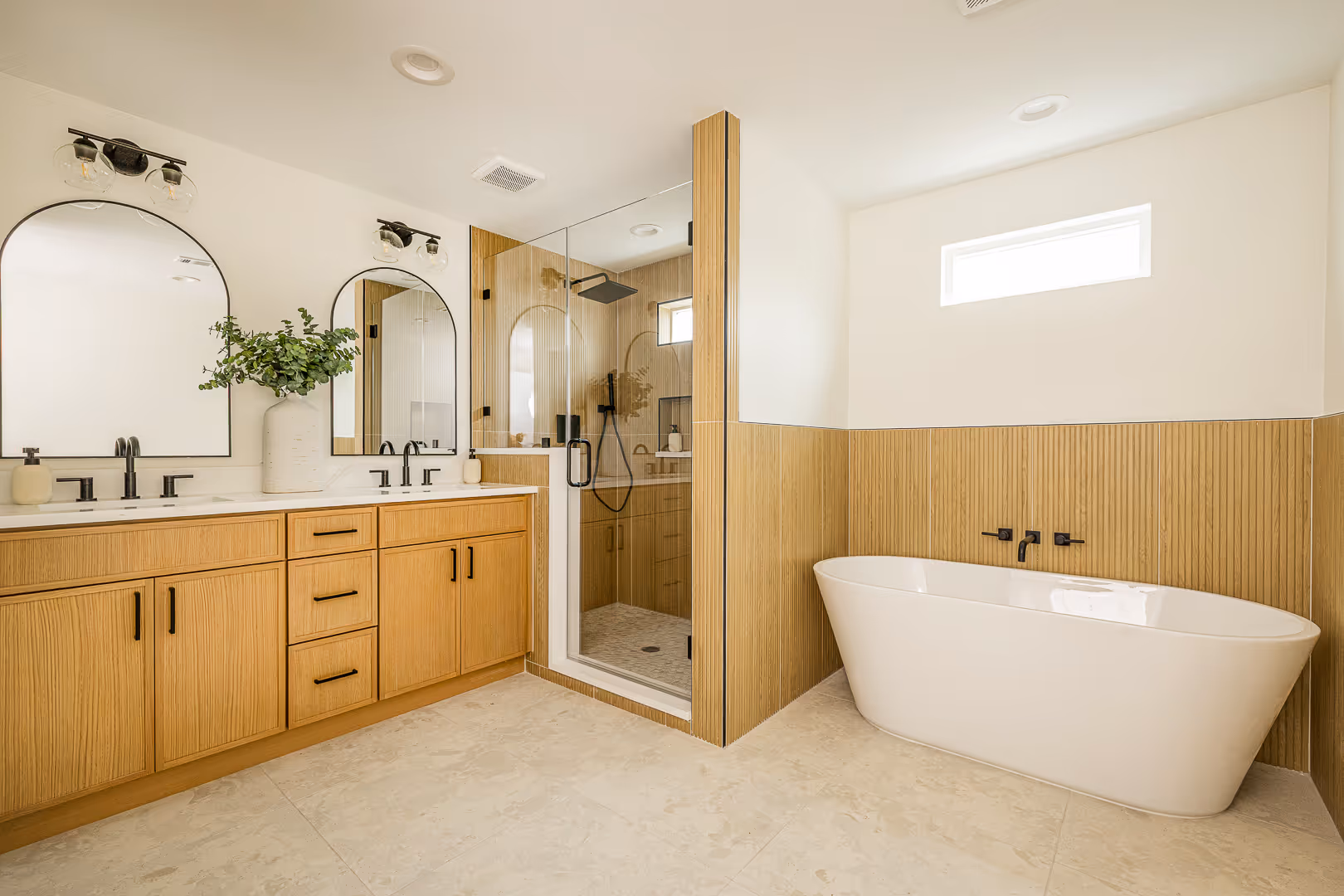Modern bathroom with a double vanity featuring light wood cabinets and arched mirrors, a glass-enclosed shower, and a freestanding white bathtub against a wood-paneled wall.