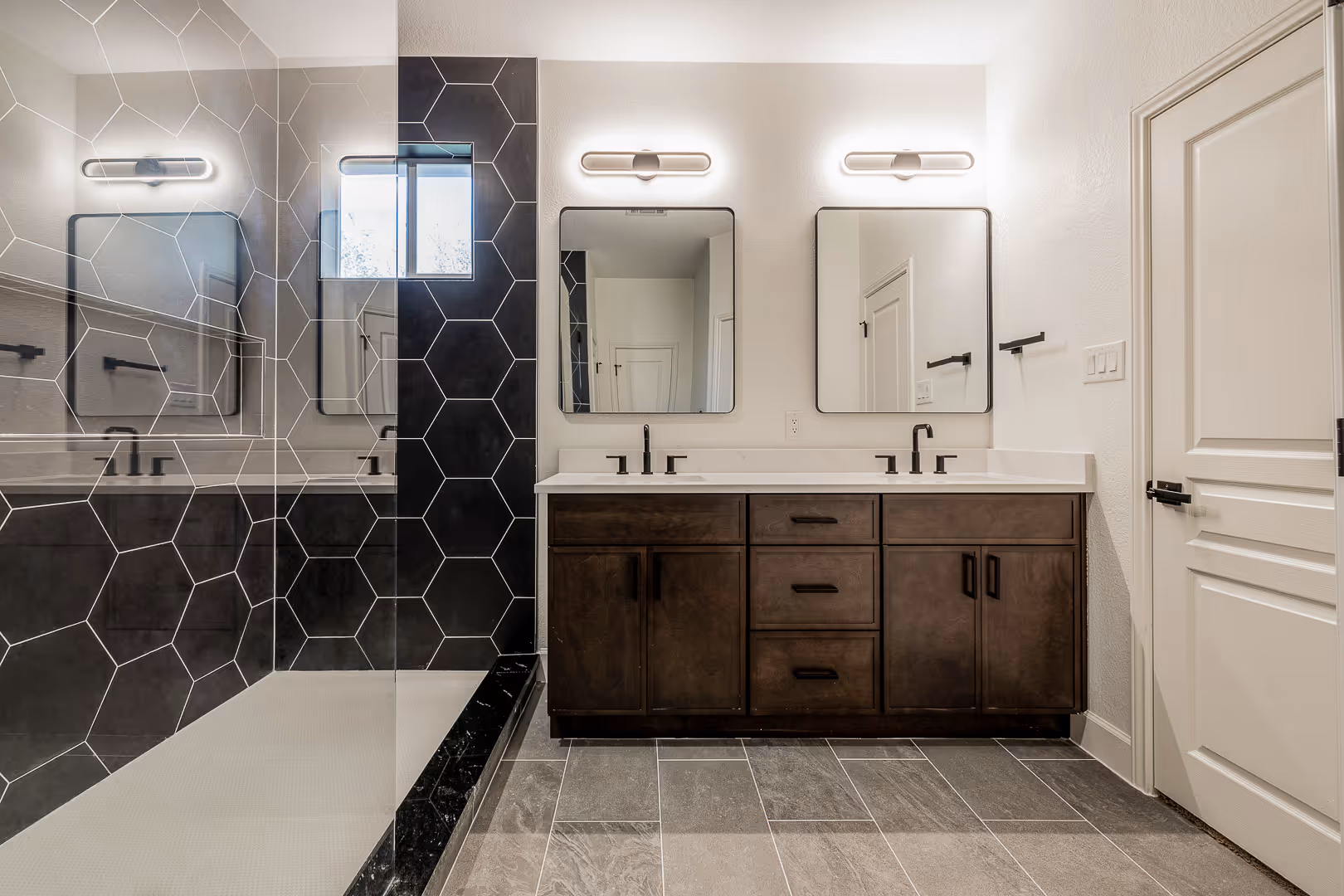 Modern bathroom with a double sink vanity featuring dark wood cabinets, two rectangular mirrors, and a walk-in shower with black hexagonal tile and glass door.