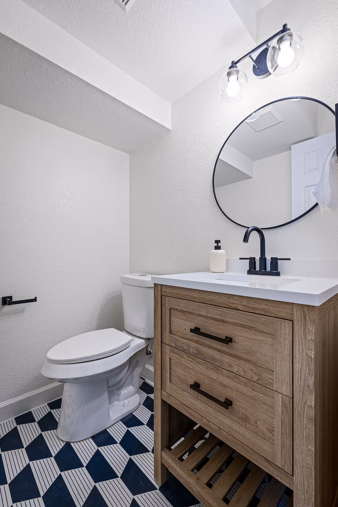 Modern bathroom design with a white toilet, wooden vanity with black faucet, round mirror, and geometric patterned floor tiles, Austin.