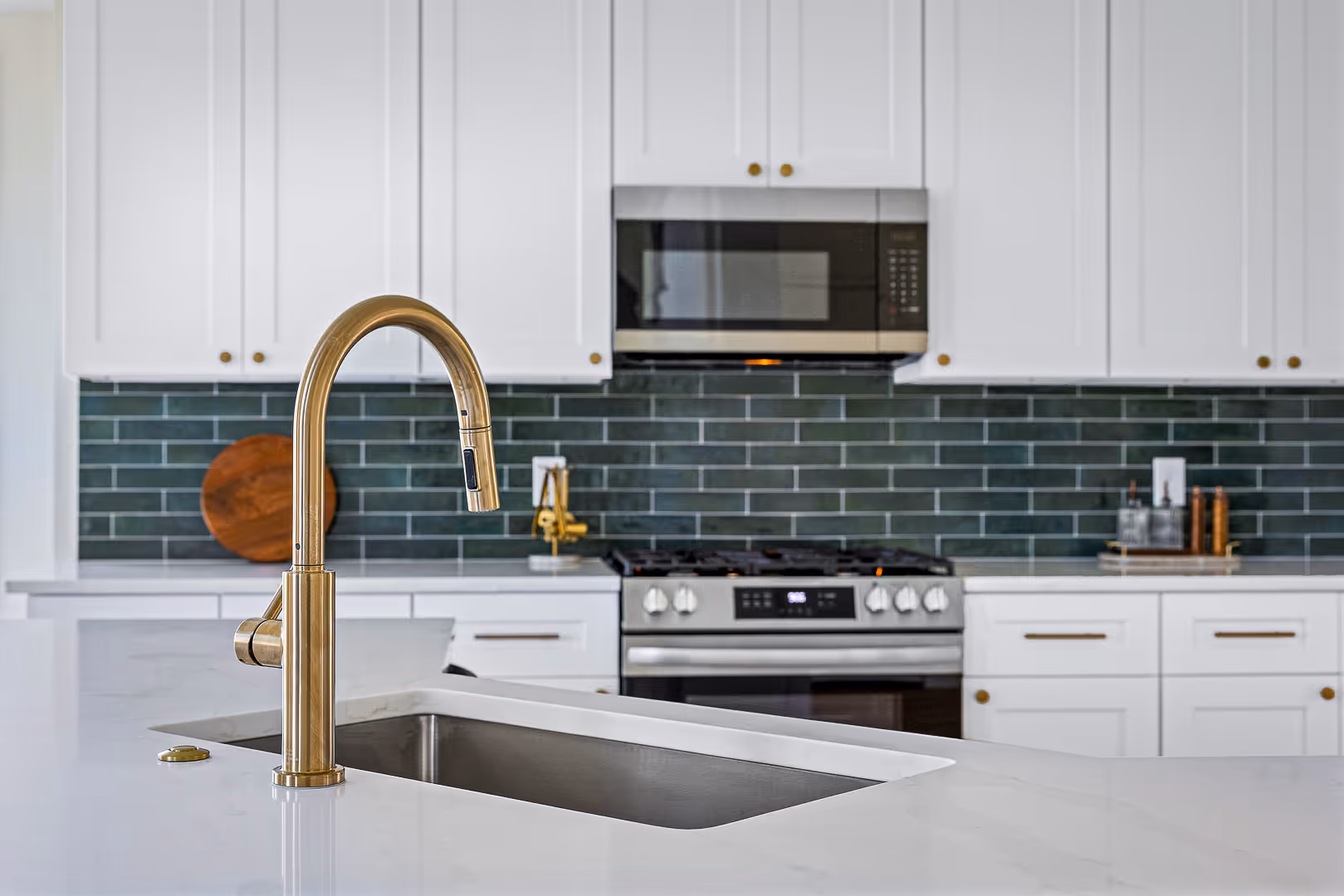 Modern kitchen design with white cabinets, green subway tile backsplash, stainless steel oven and microwave, and a gold faucet on a white countertop island with an undermount sink.