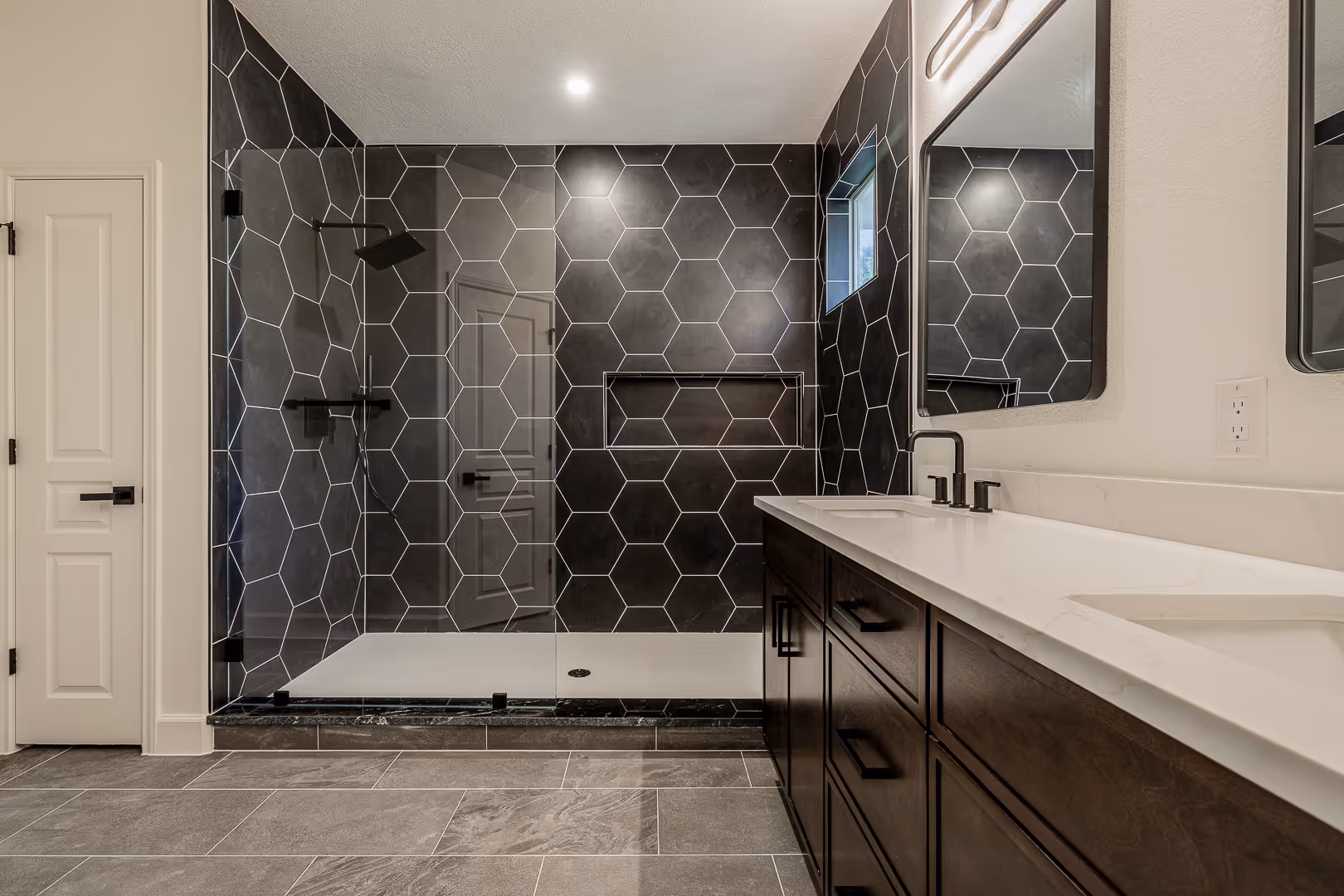Modern bathroom with large walk-in shower featuring black hexagonal tiles and white grout, dual sinks with dark wood cabinetry, and a rectangular mirror.