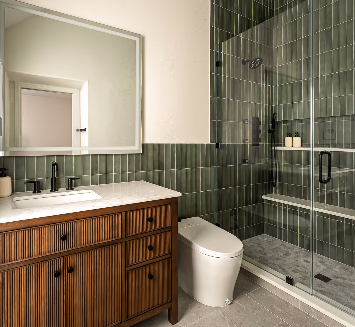 Modern bathroom with wood vanity, white marble countertop, white toilet, and glass shower with green vertical tile walls.