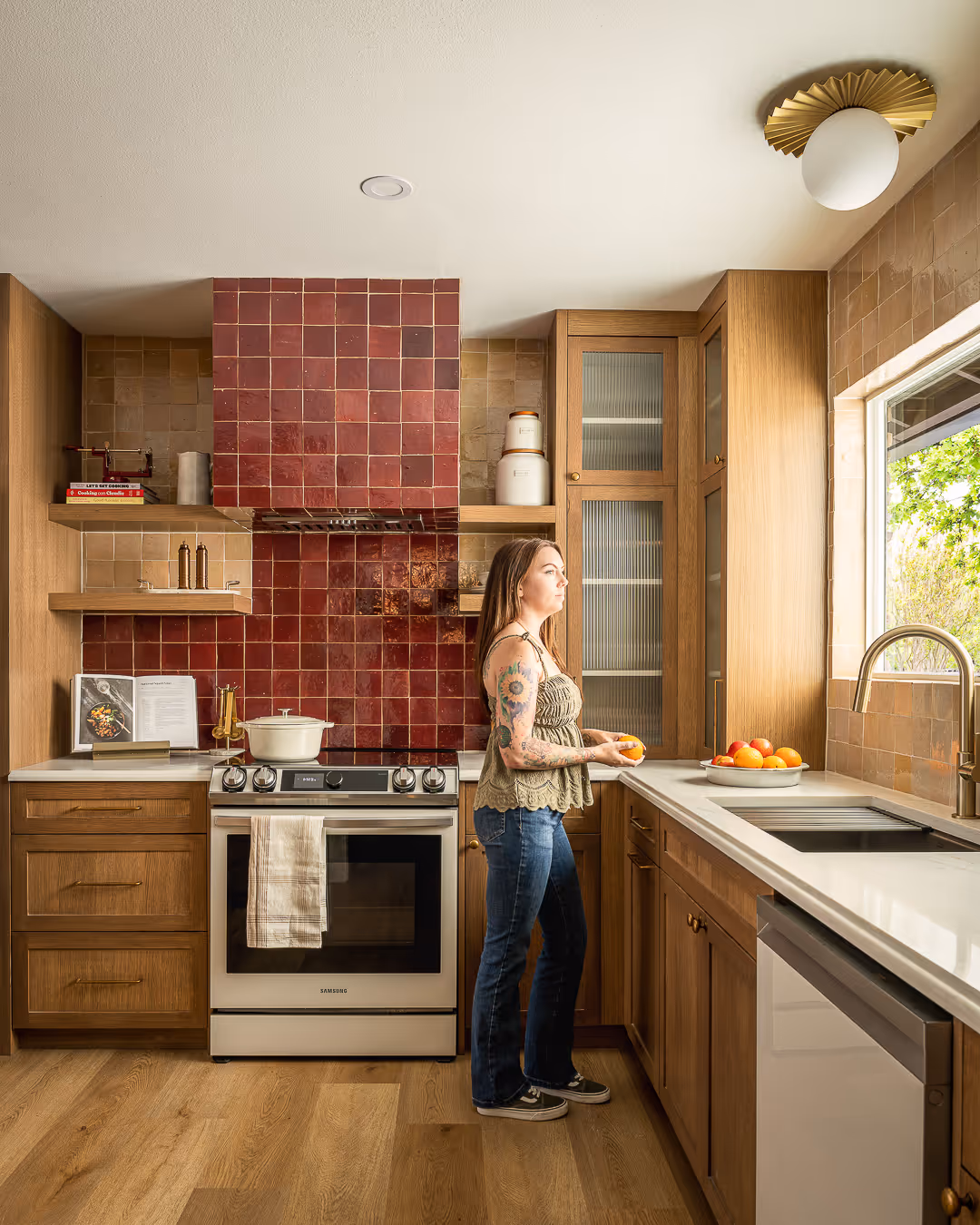 Woman standing in a modern kitchen with wooden cabinetry, red tiled backsplash, and holding an orange by the countertop near a window with a fruit bowl.