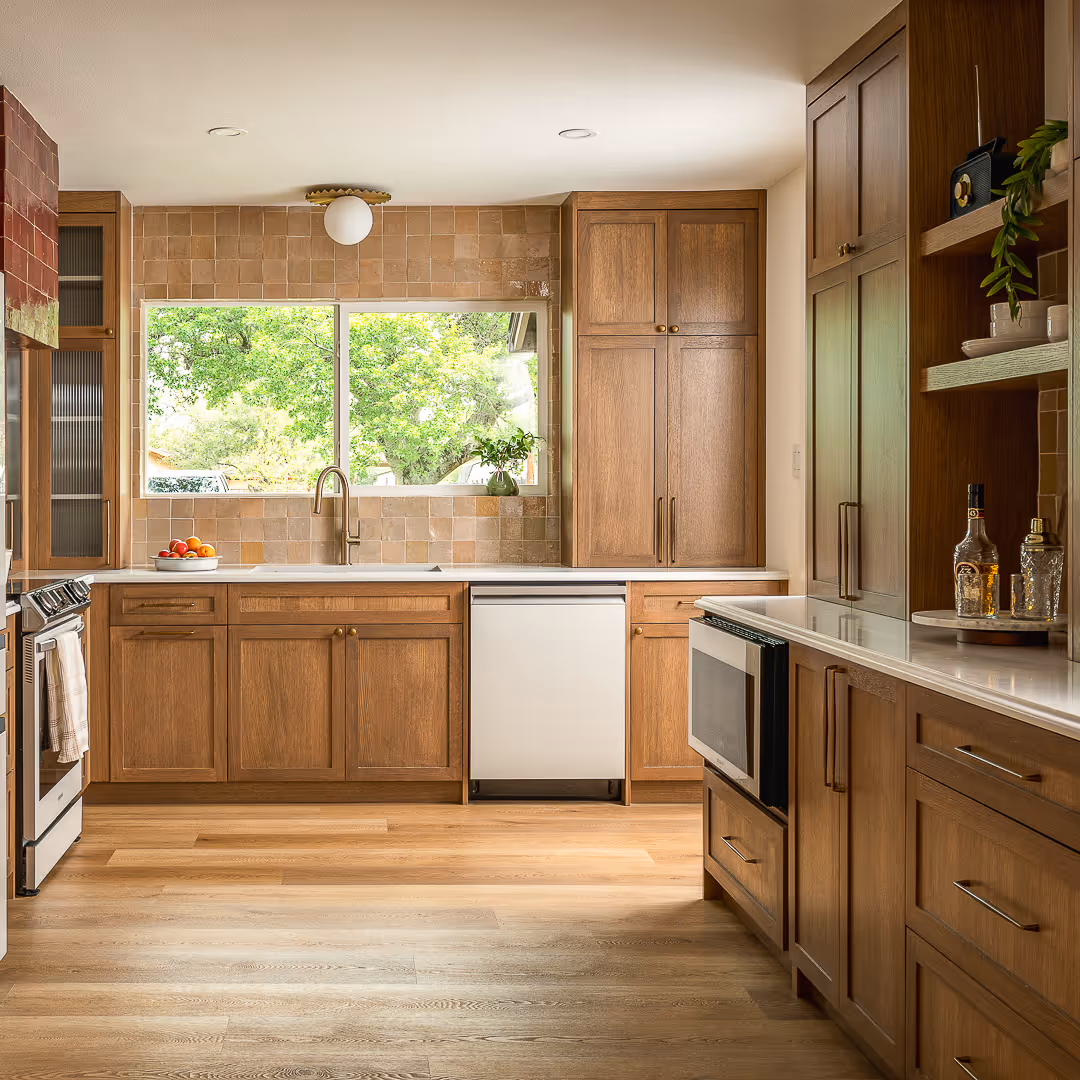 Modern kitchen with wood cabinets, light tile backsplash, white countertops, a window above the sink showing green trees outside, and stainless steel appliances.