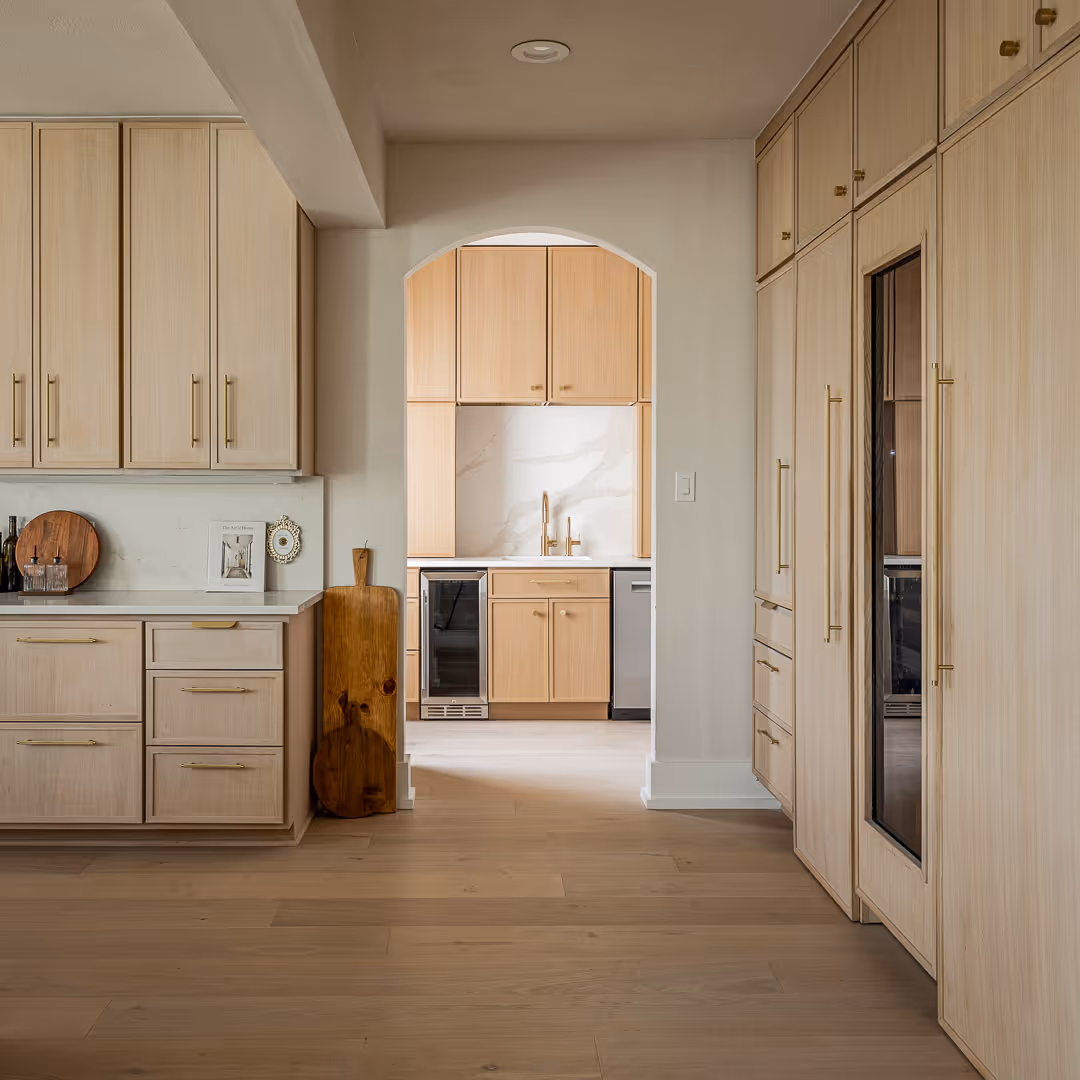 Remodeled modern kitchen with light wood cabinetry, gold handles, white countertops, and an arched doorway leading to a second kitchen area with a sink and wine cooler.