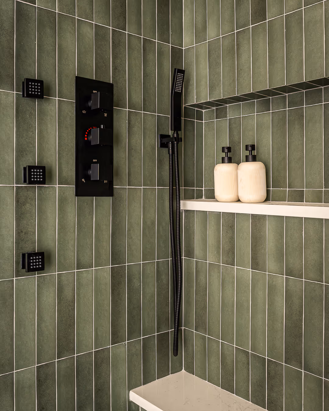 Shower corner with vertical green tiles, black shower controls, black handheld showerhead, and a white shelf holding two beige pump bottles.