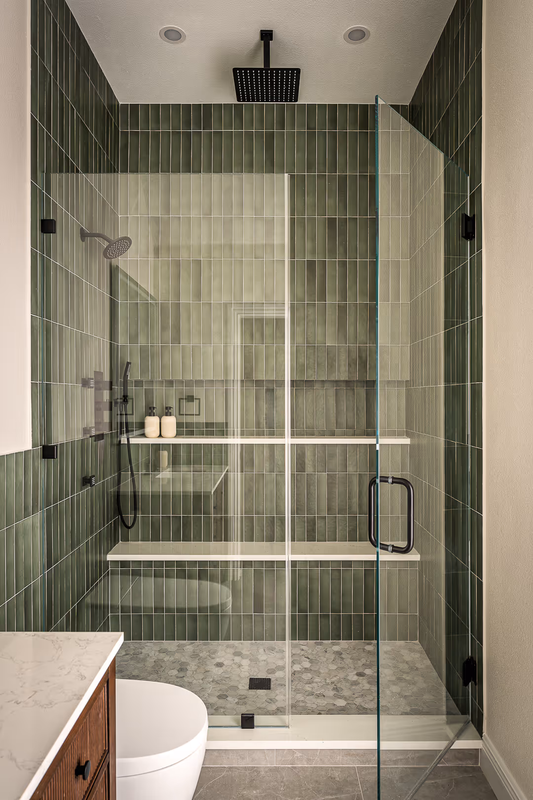 Modern bathroom shower with green vertical tiles, glass door, black rain shower head, and hexagonal gray floor tiles.
