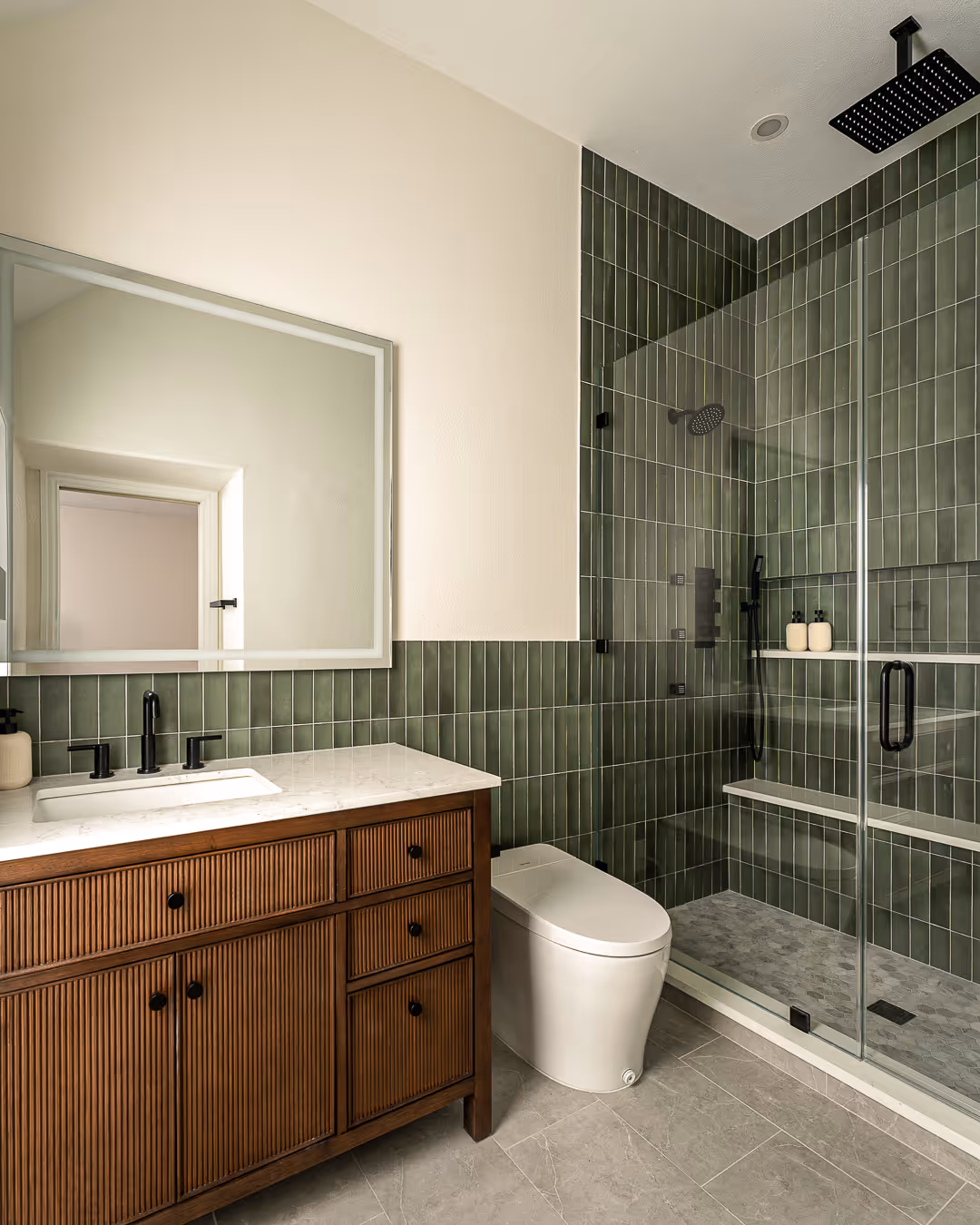 Modern bathroom with green vertical tile walls, wooden vanity with white countertop, wall-mounted mirror, white toilet, and glass-enclosed shower with black fixtures.
