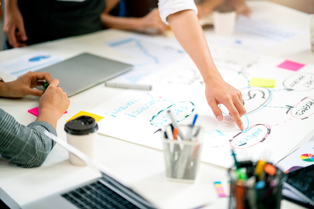 People collaborating around a table with charts, notes, laptops, and coffee cups during a brainstorming session.