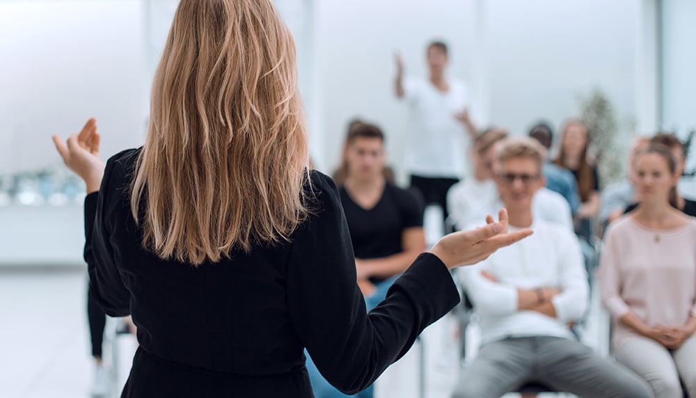 Woman with long blonde hair in black jacket speaking to a seated audience in a bright room.