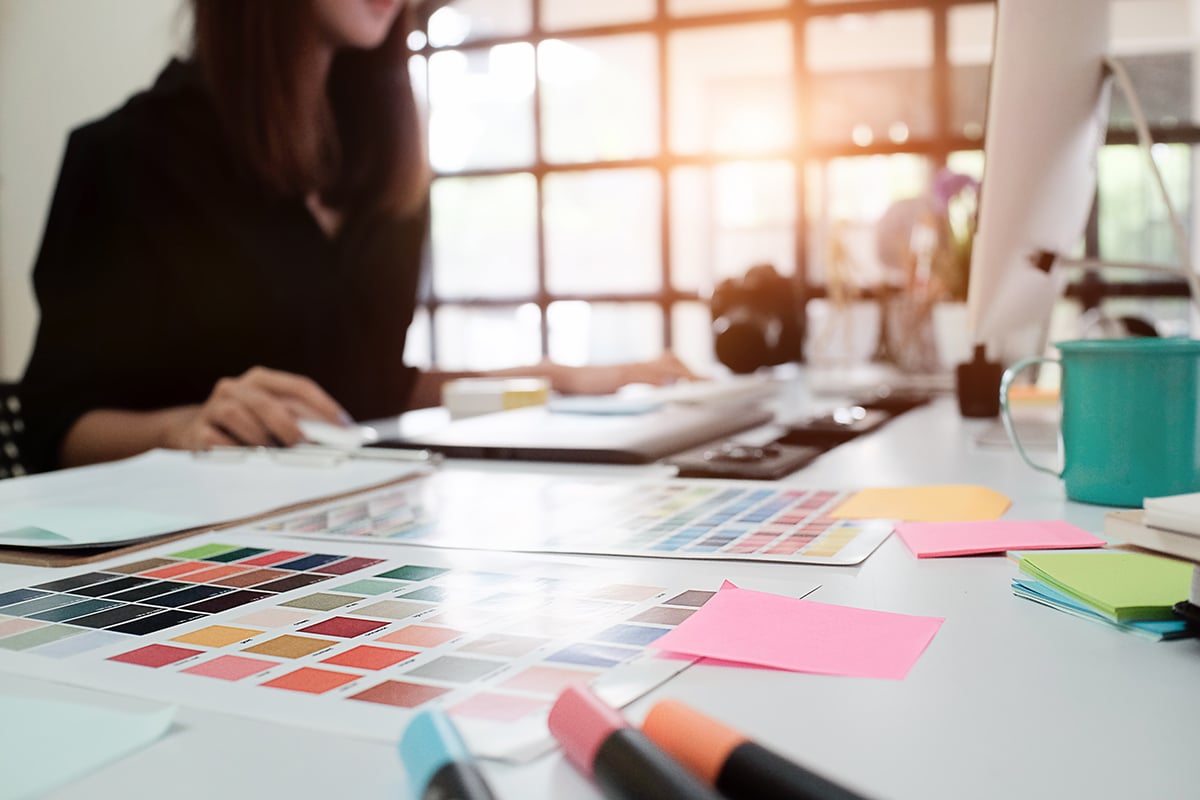 Designer working at a desk with color swatches, sticky notes, markers, and a computer in a bright office.