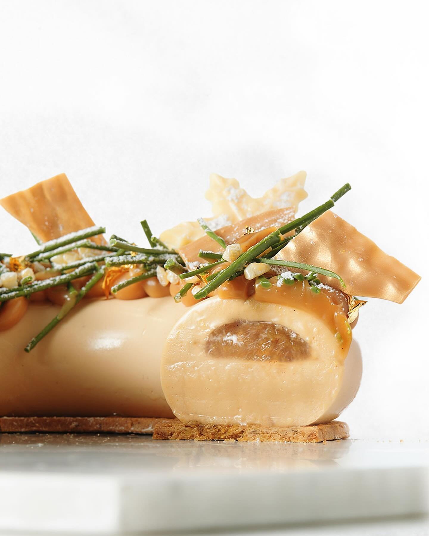 Close-up of a beige mousse dessert log with caramel topping, green sprigs, white edible decorations, and a crumbly base.