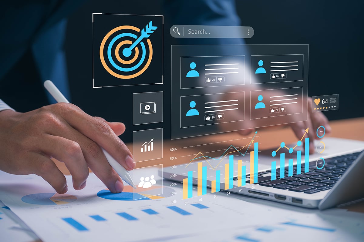 Person using a stylus pen on financial charts with digital interface showing graphs, social media profiles, and a target icon above a laptop keyboard.