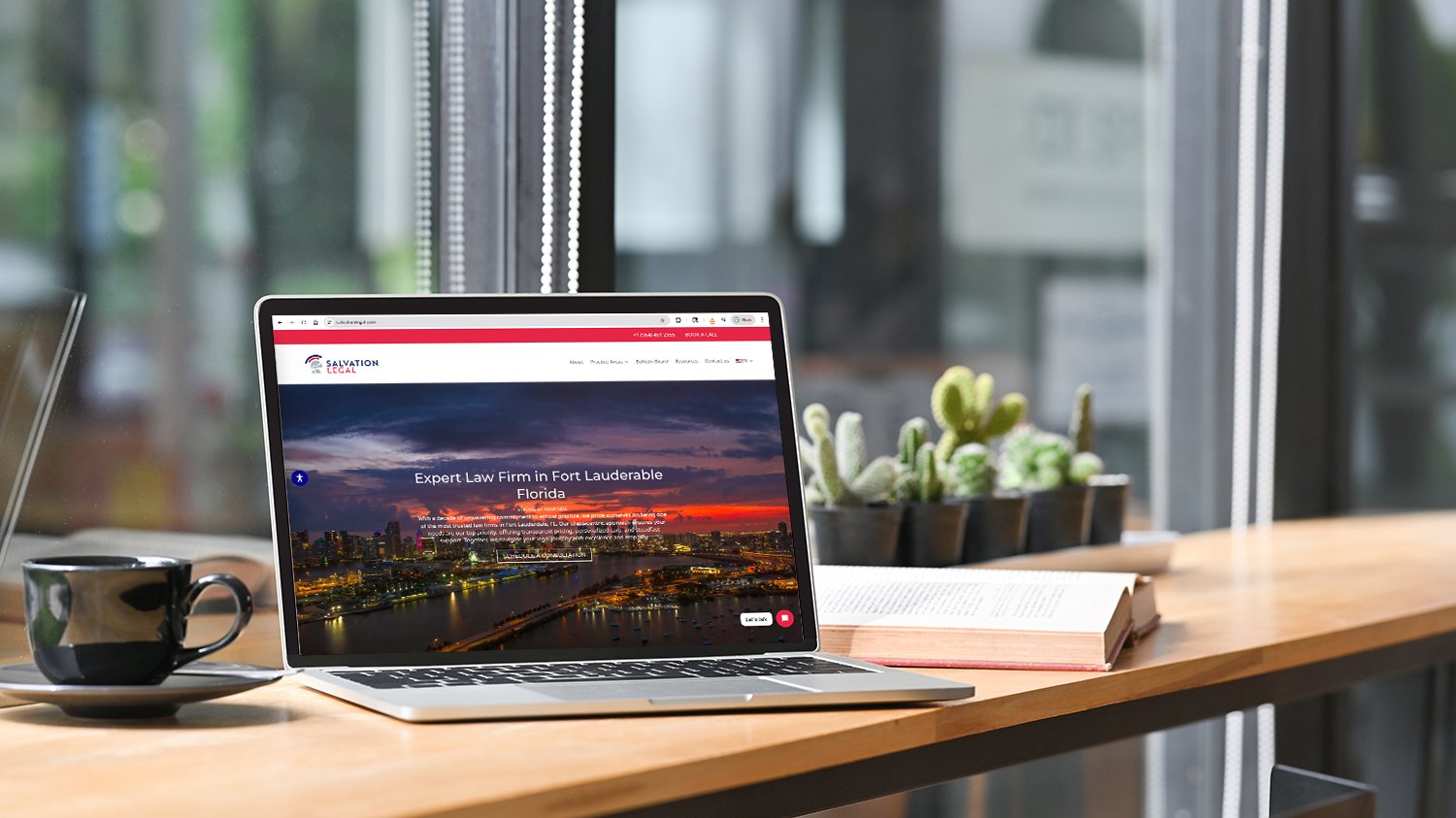 Laptop on a wooden table showing a law firm website with a cityscape background, next to a black coffee cup and an open book with small potted cacti behind.