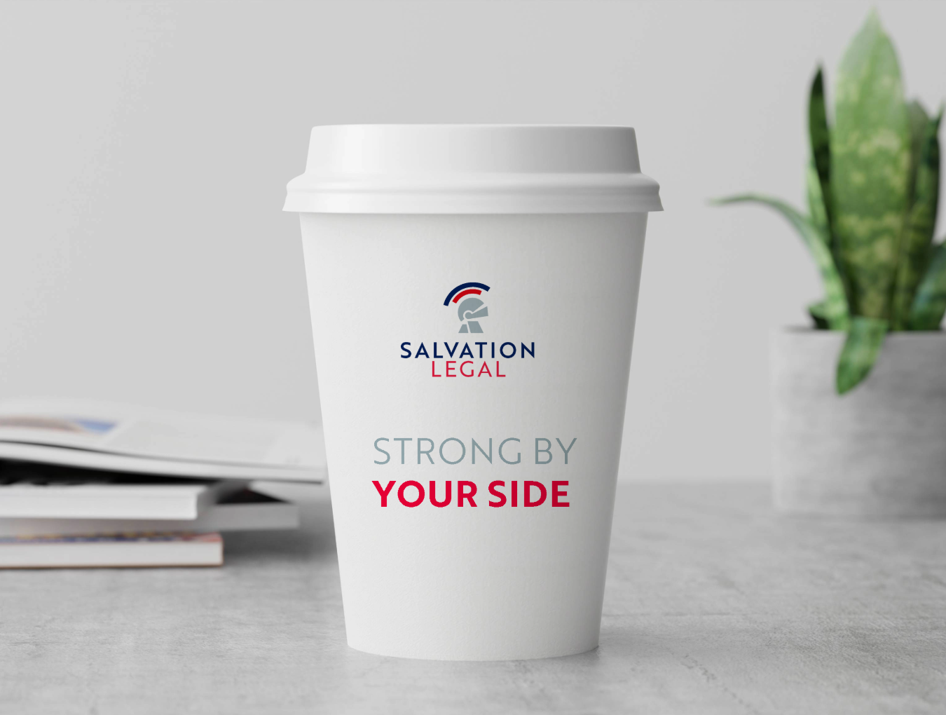 White disposable coffee cup with a white lid and Salvation Legal logo, text reads 'Strong by Your Side', placed on a light grey surface with blurred books and a plant in the background.