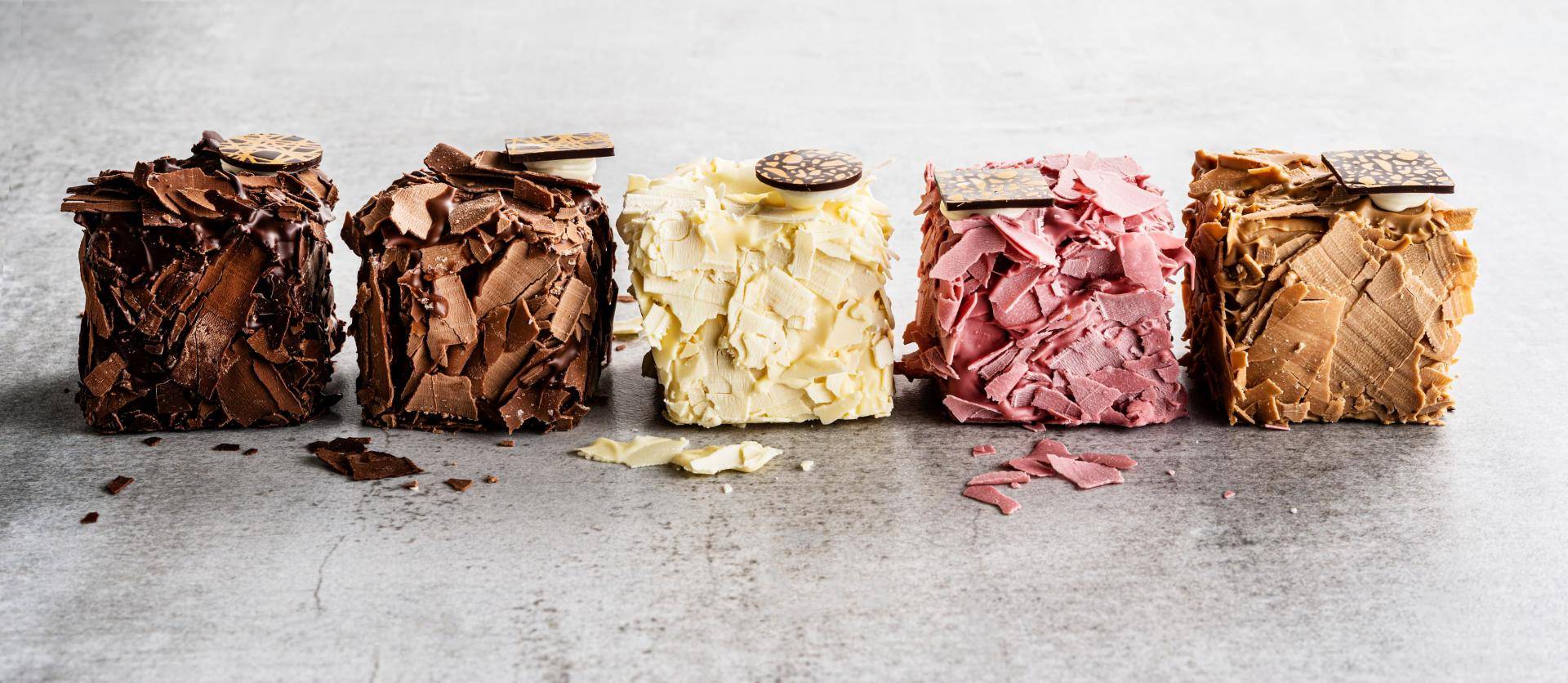 Five square chocolate desserts covered with dark, milk, white, raspberry, and caramel chocolate shavings, each topped with a decorative chocolate disc.