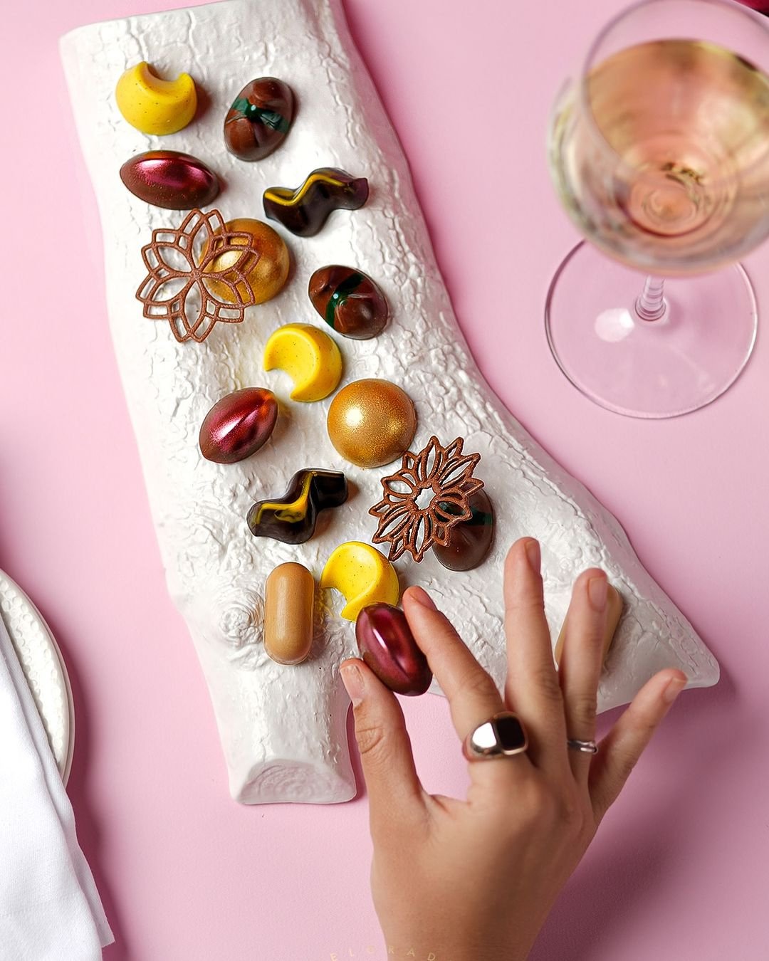 Hand picking up a shiny, oval chocolate from a white textured serving platter with various colorful chocolates and a glass of white wine on a pink surface.