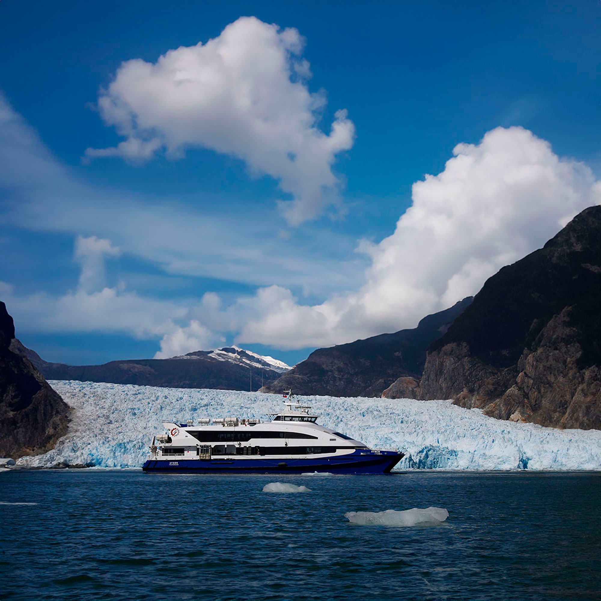 White and blue passenger ferry sailing on water with glacier and mountains in the background under a partly cloudy sky.