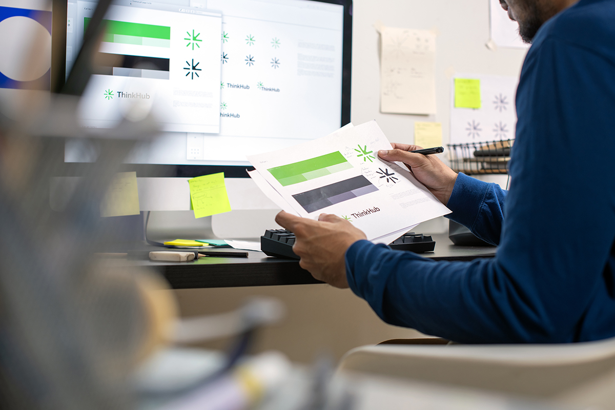 Person holding printed branding designs with green and black color palettes labeled ThinkHub, reviewing them in front of a computer screen showing the same designs.