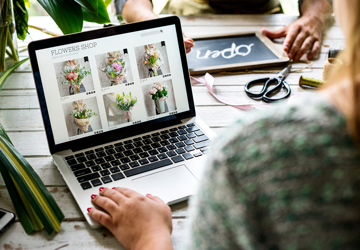 Person browsing an online flower shop on a laptop with images of various bouquets, while another person holds a chalkboard sign that says 'open' on a rustic wooden table.