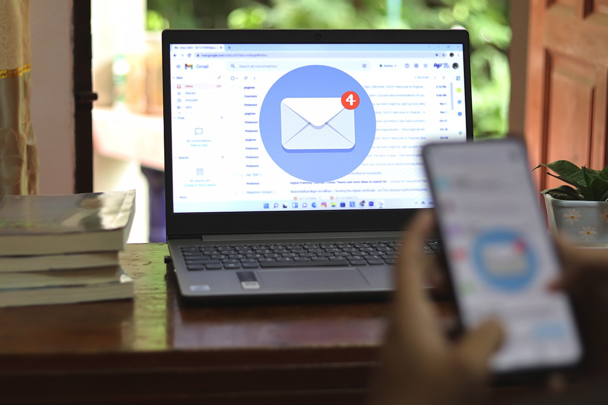 Laptop on a wooden table showing Gmail inbox with a notification of 4 new emails; blurred hands holding a phone in the foreground.