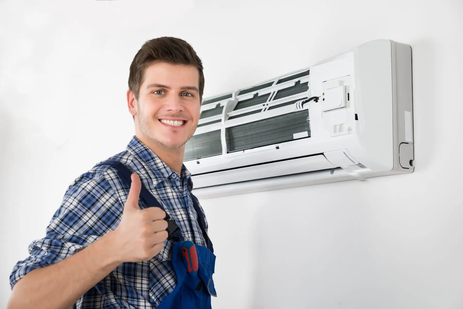 A smiling technician in blue overalls is giving a thumbs-up while standing next to a mini-split unit