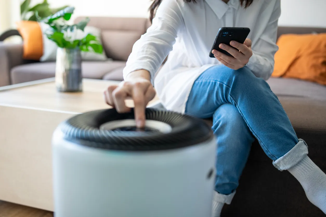 A person sits on a sofa looking at their phone next to an air purifier
