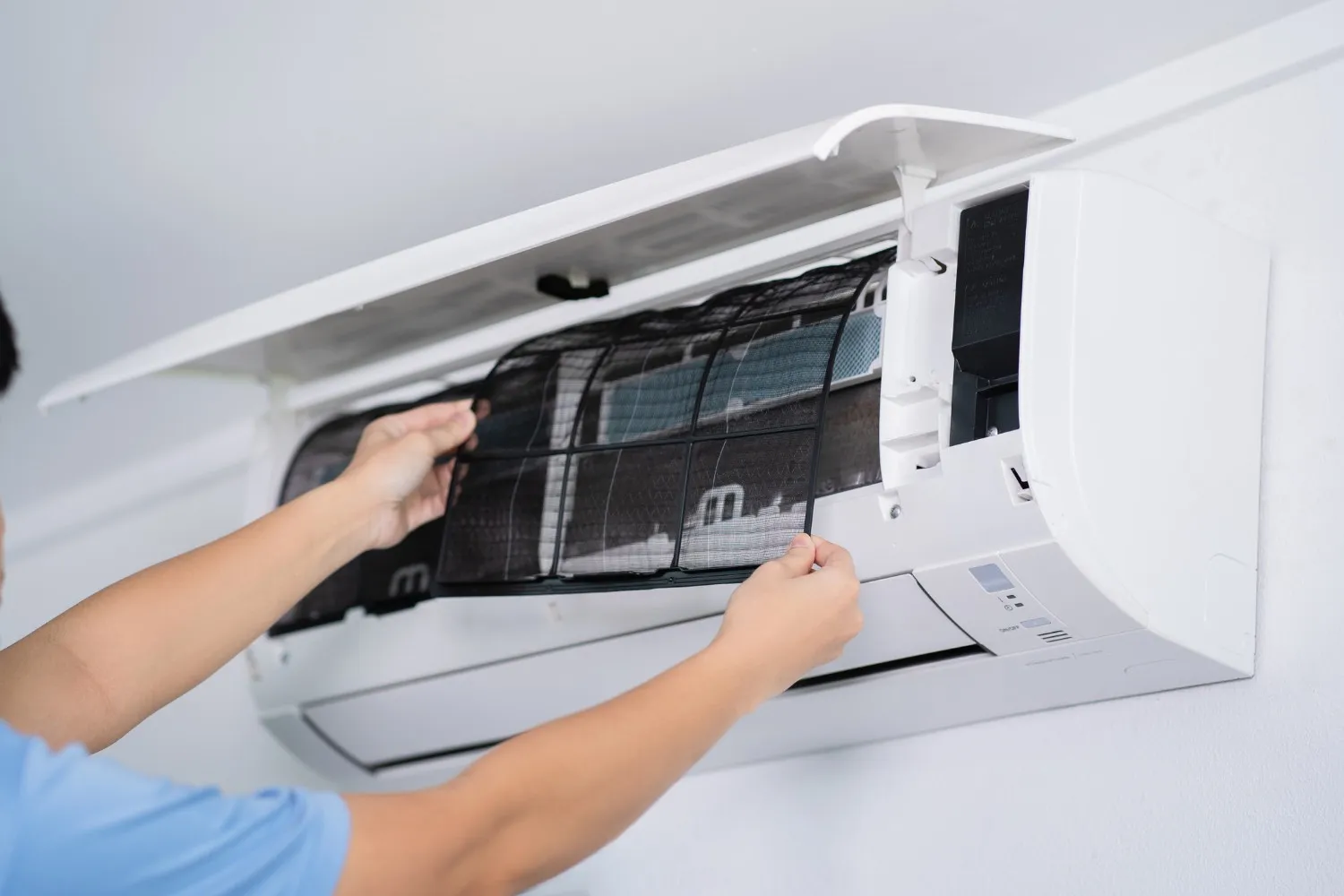 A person in a blue shirt removes a black mesh dust filter from a wall-mounted white air conditioning unit. The front cover of the AC unit is propped open to allow access to the internal components.