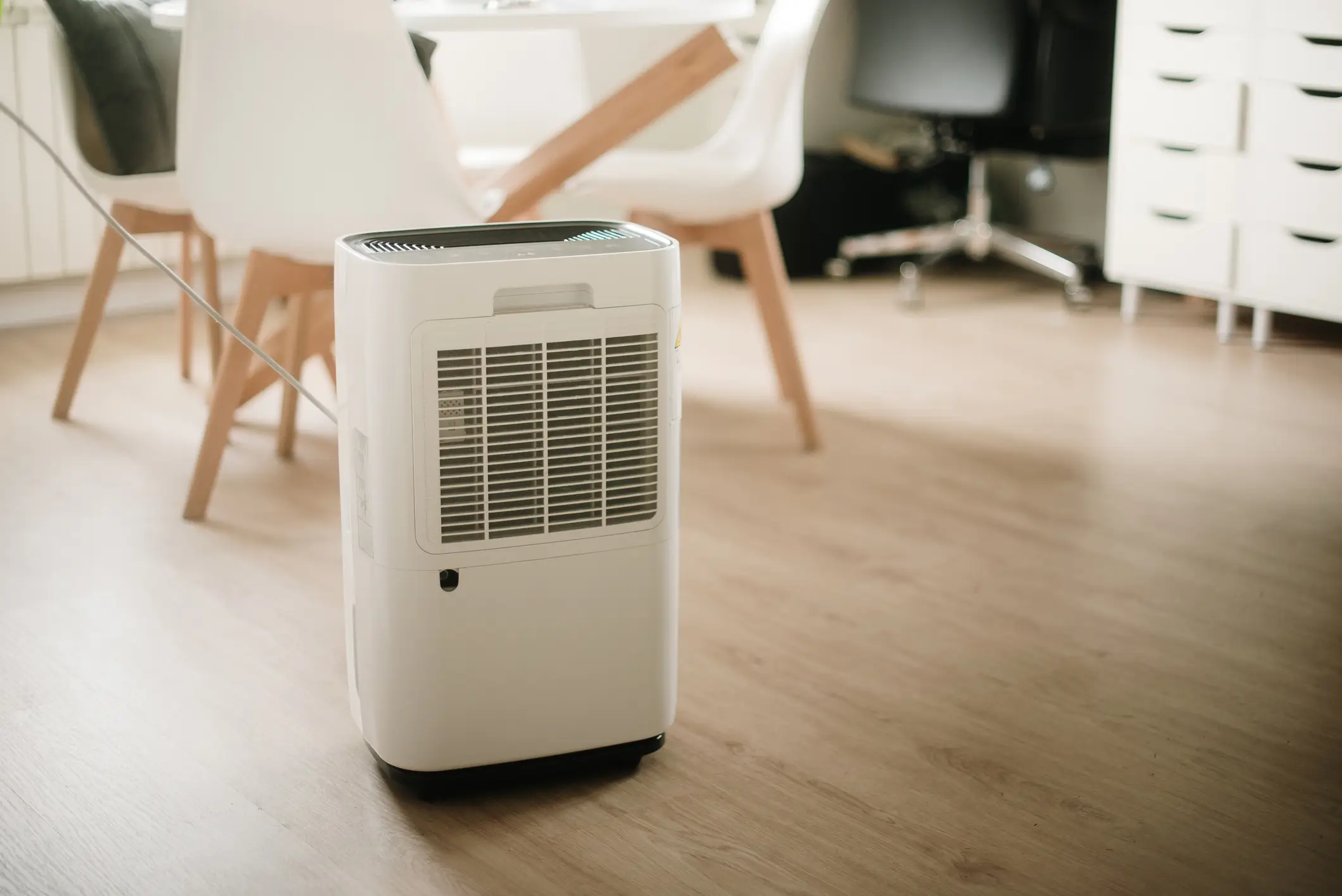 A white portable air purifier or dehumidifier stands on a light-colored wood floor in a brightly lit dining area. It features a large front intake grille and a top-mounted control panel.