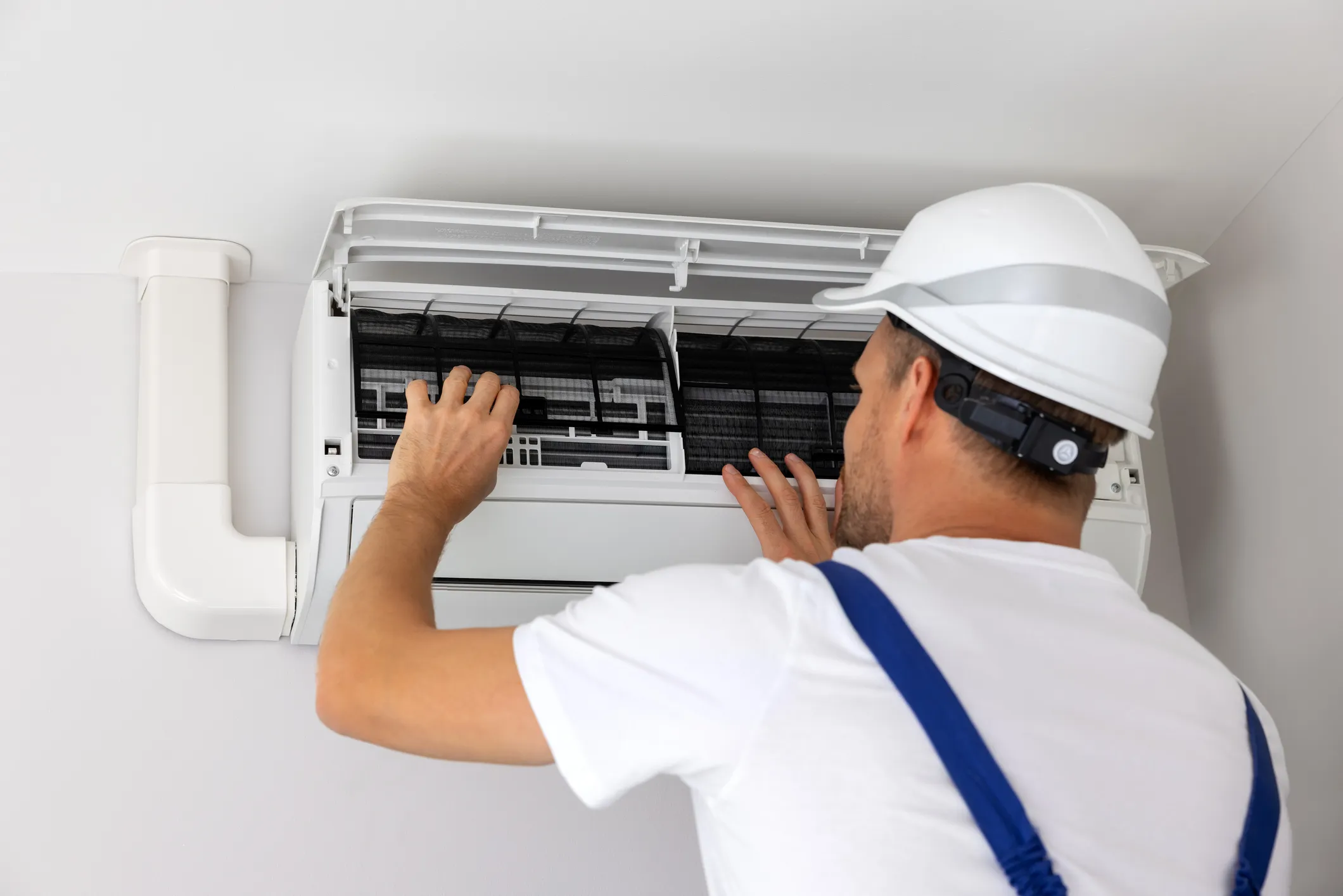 HVAC technician in a hard hat cleaning or checking the air filters of an indoor AC unit.