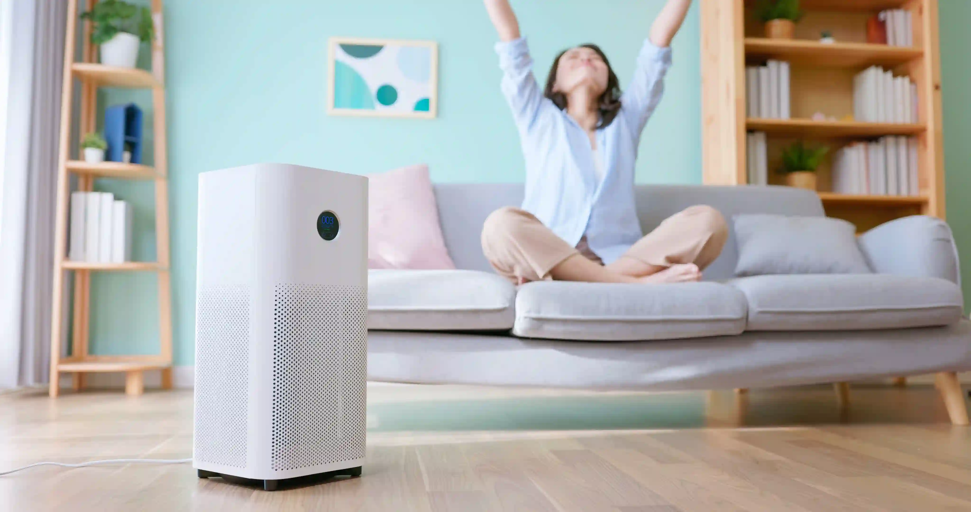 A white rectangular air purifier with a circular digital display stands on a wooden floor in a living room, while a woman sits on a grey sofa in the background with her arms raised in a stretch.