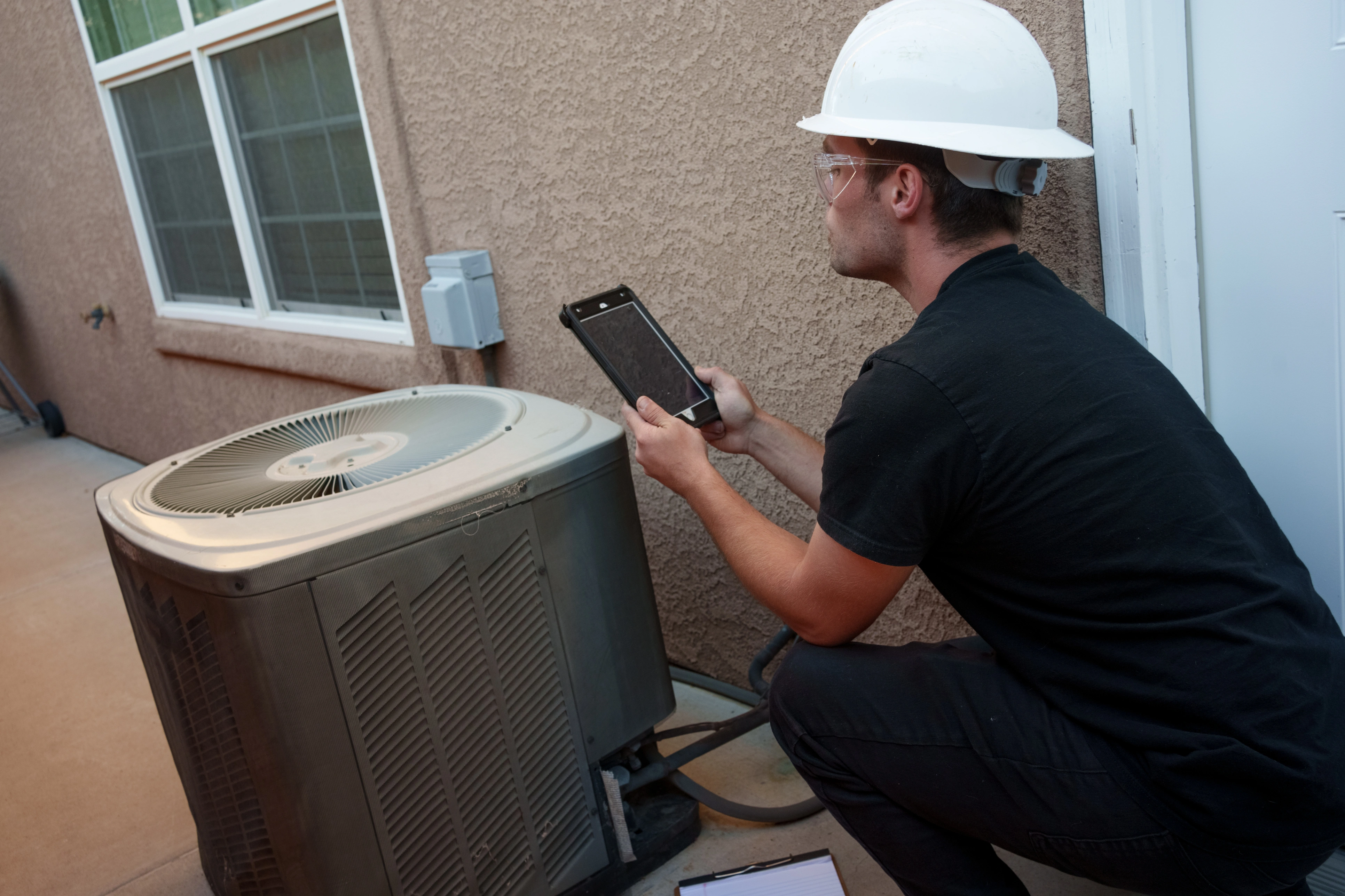 A technician in a white hard hat crouches beside an outdoor air conditioning unit