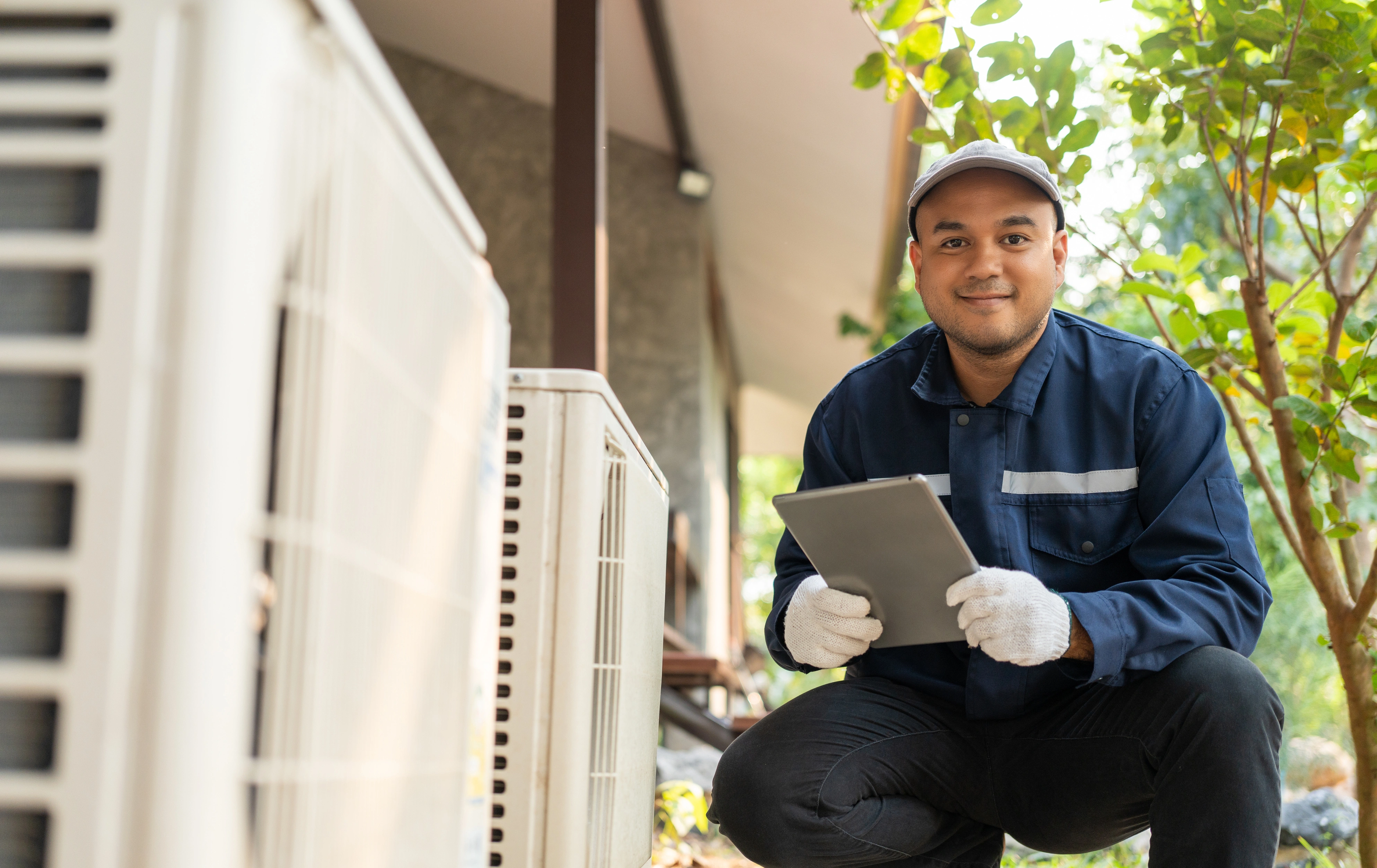 A technician in a blue uniform kneels outside beside an HVAC unit, holding a tablet.