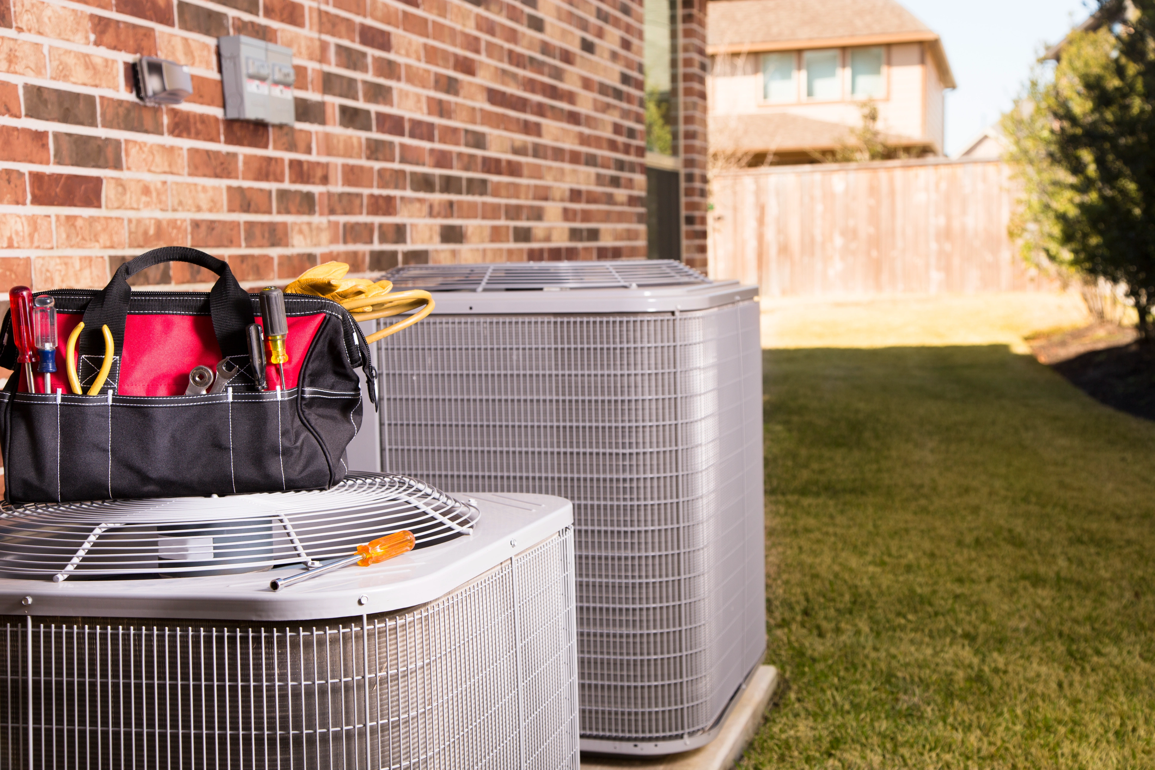 HVAC technician tool bag resting on outdoor air conditioning condenser units beside a brick home