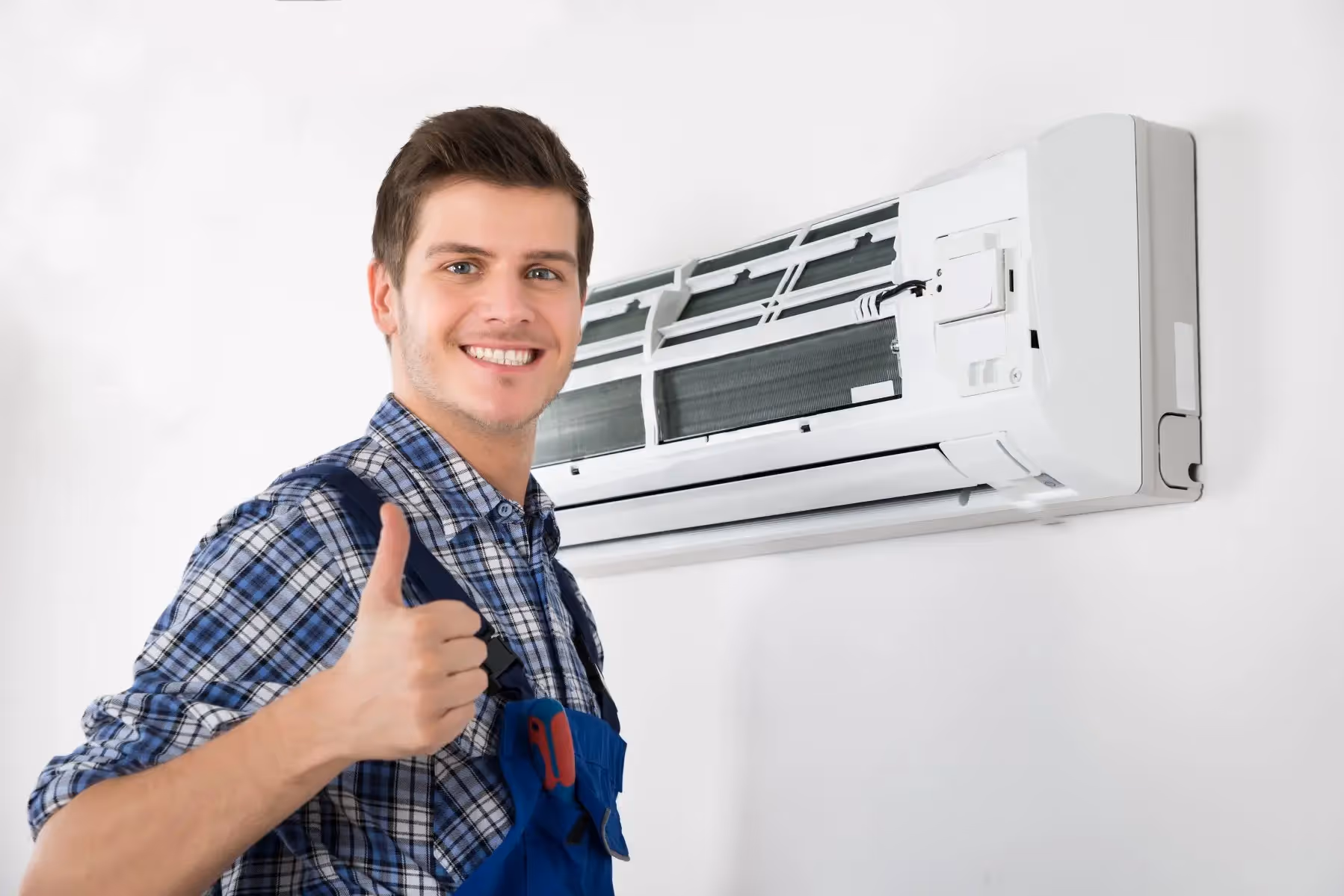 A smiling technician in blue overalls is giving a thumbs-up while standing next to a mini-split unit