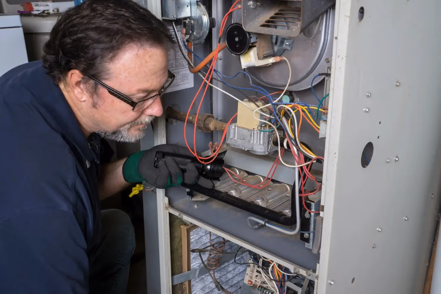 A technician inspects the burner assembly and wiring inside a residential forced-air furnace.