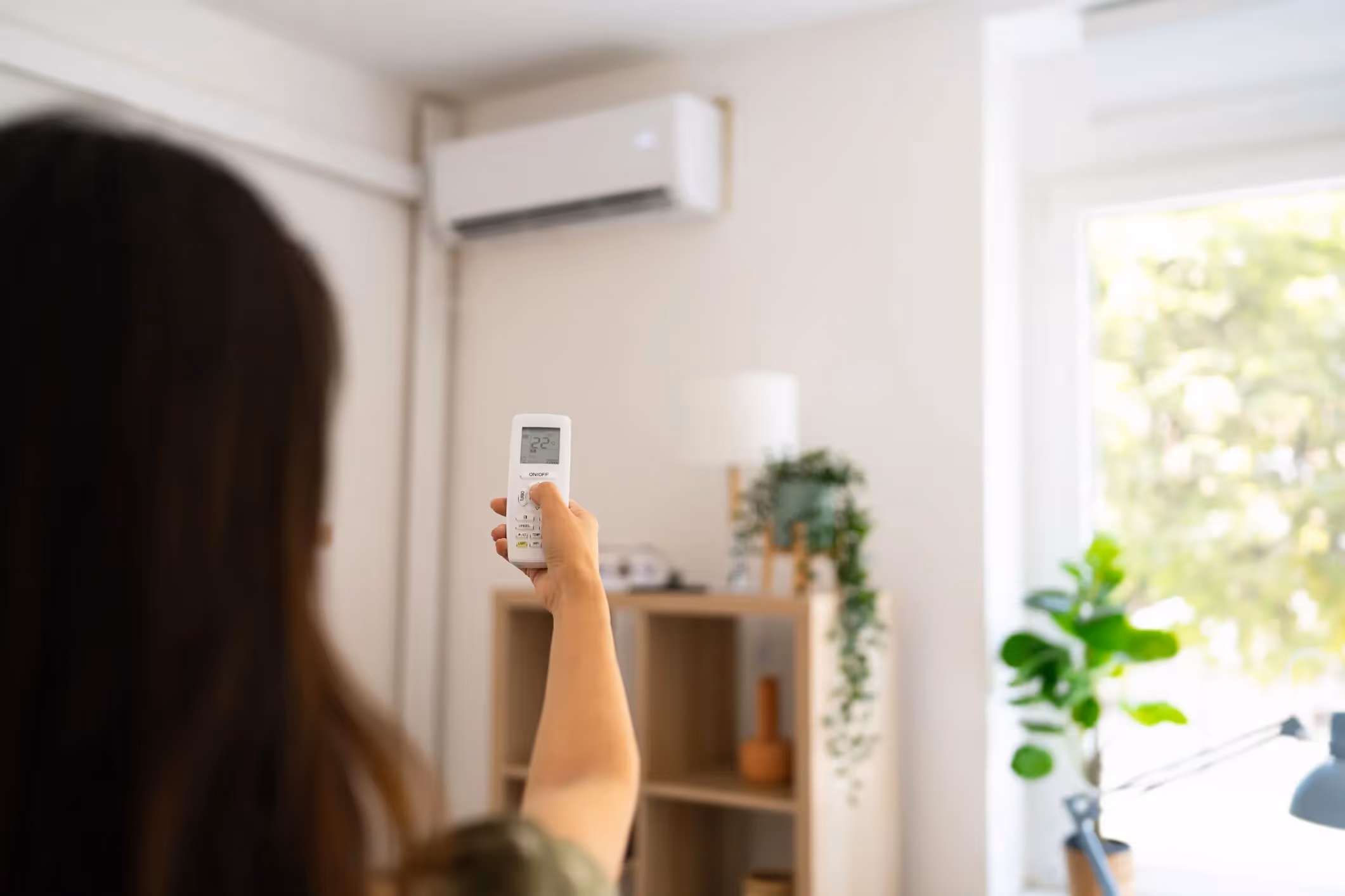 A person is holding an AC remote, pointing it at a wall-mounted air conditioning unit to adjust the temperature.