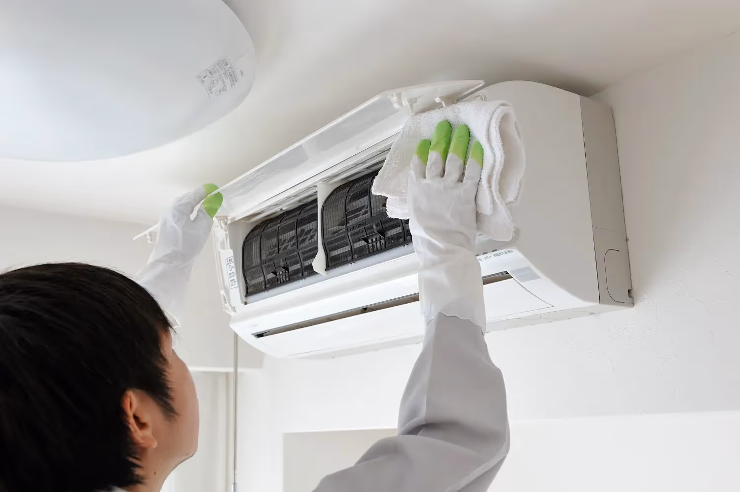 A person wearing protective gloves cleans a wall-mounted air conditioning unit with a white towel. The unit's front cover is open, exposing the internal air filters for maintenance against a white wall.