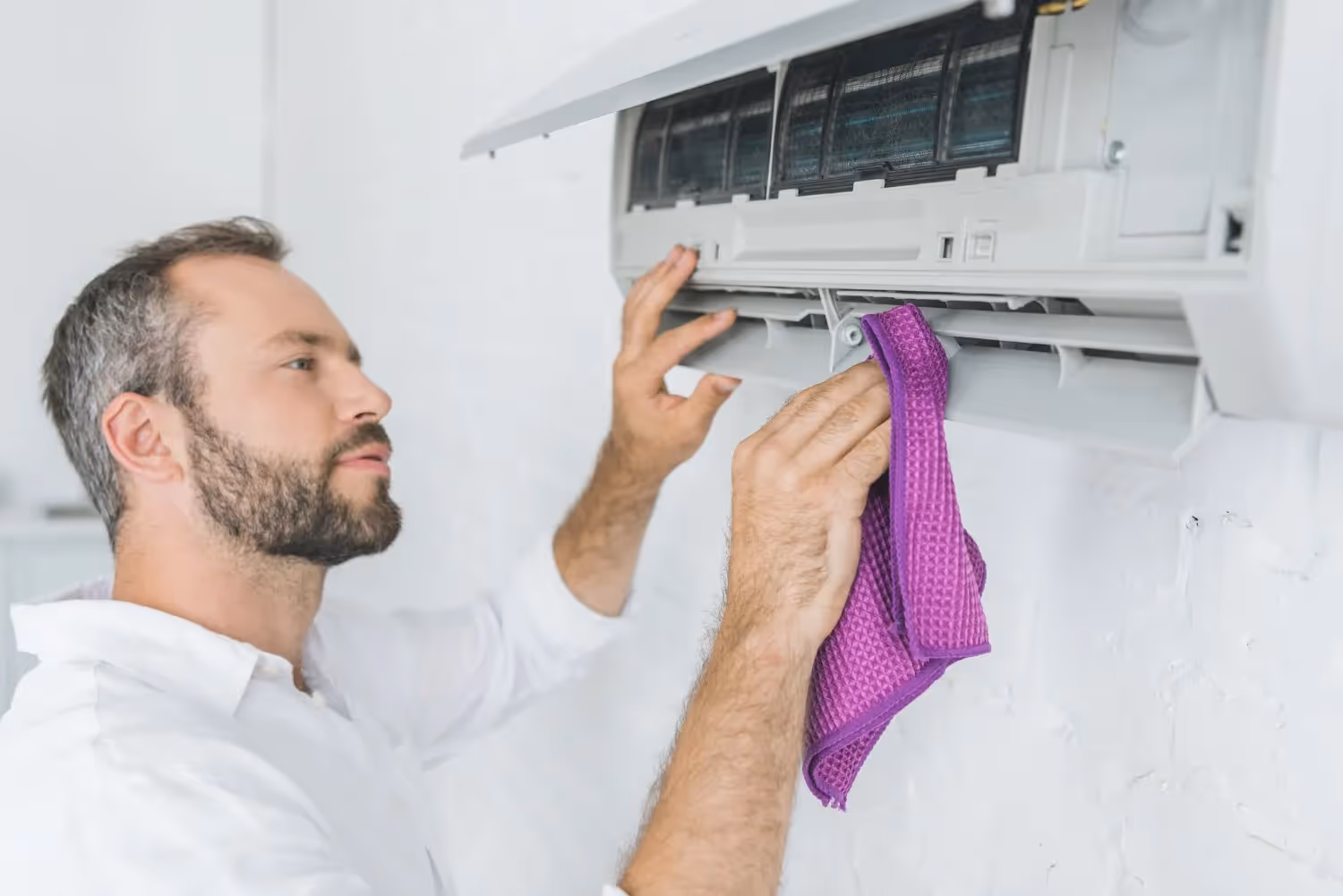 A man wipes the vents of an indoor air conditioning unit with a purple cloth. The front panel of the white AC unit is flipped open for cleaning against a white wall.