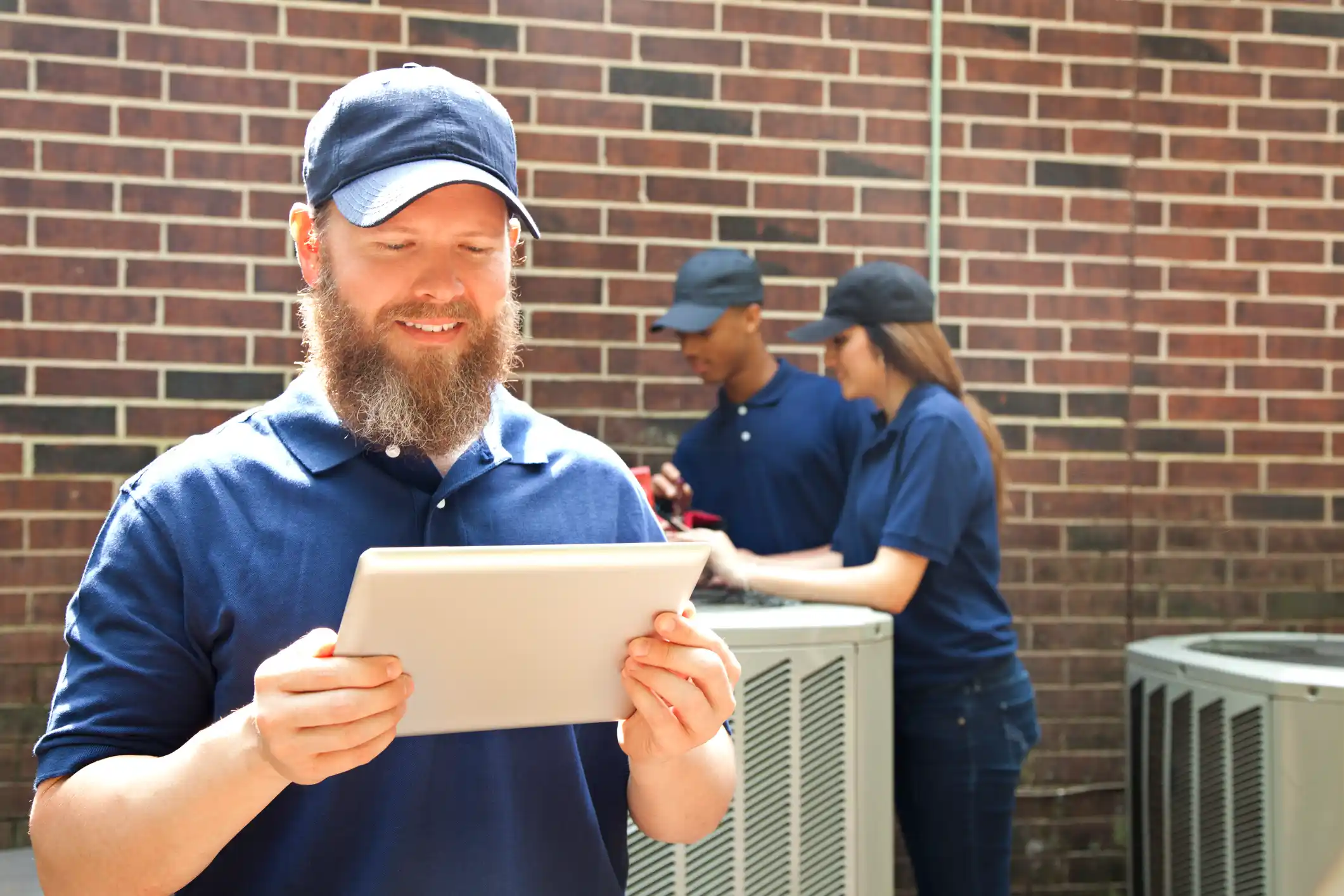 Smiling HVAC technician using a tablet while a team checks an outdoor air conditioning unit.