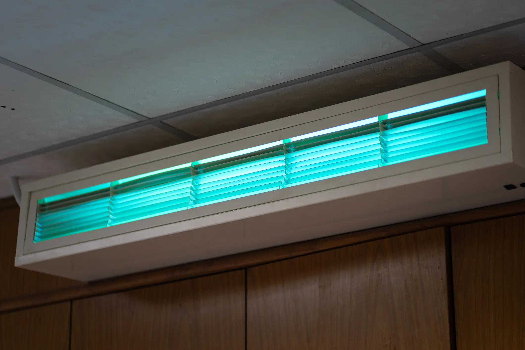 A wall-mounted UV air purifier with horizontal slats glowing with a bright turquoise light. The rectangular unit is installed near the ceiling above a wooden cabinet.