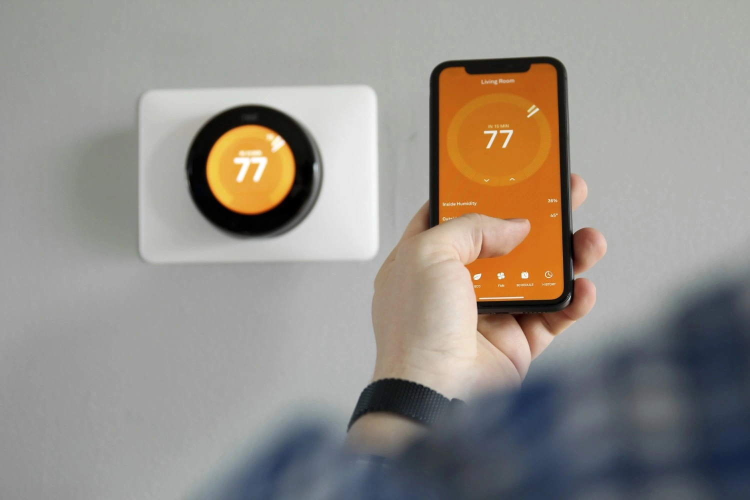 A hand holding a smartphone displaying a thermostat app next to a wall-mounted smart thermostat.