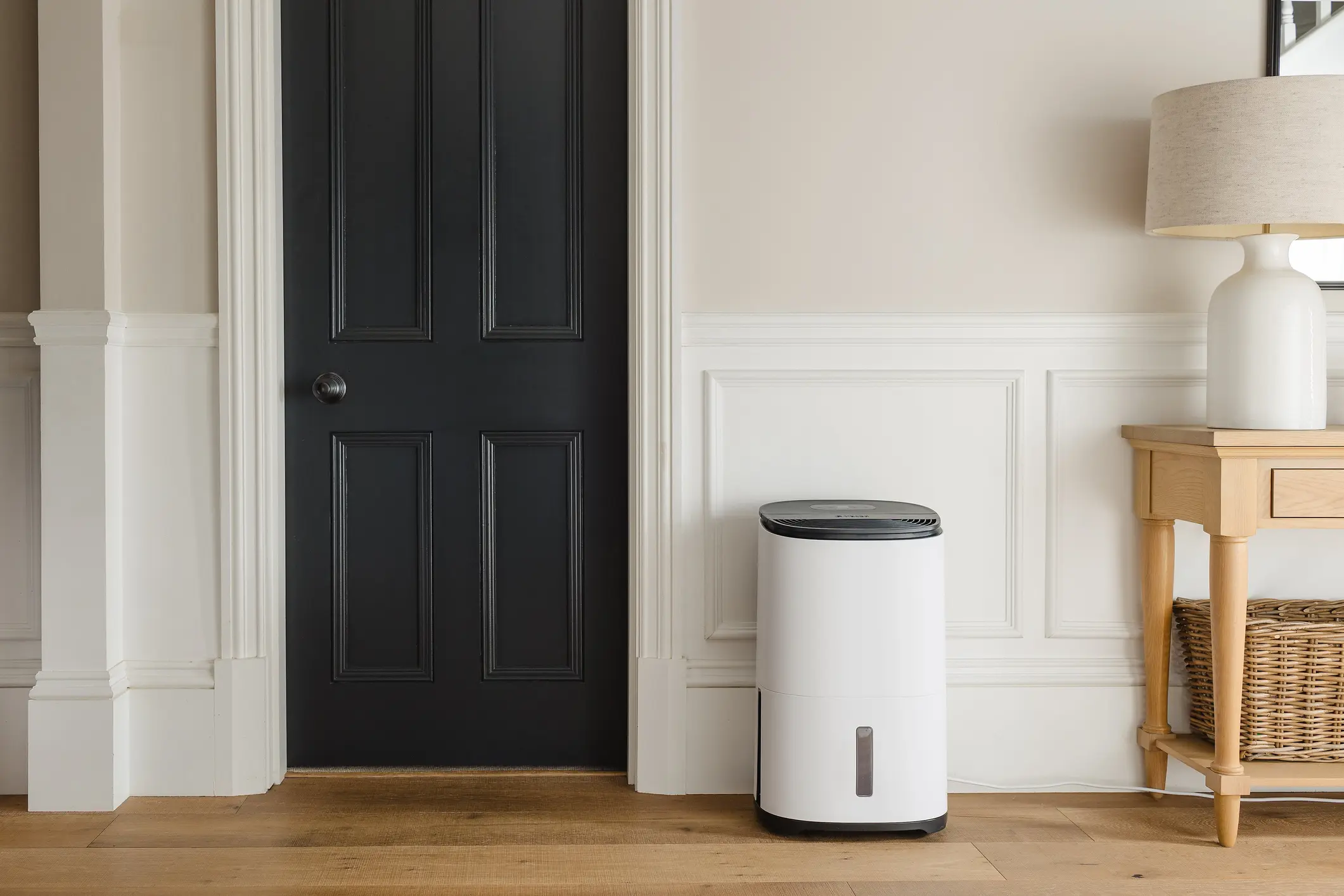A modern, cylindrical white dehumidifier with a black top is positioned on a wood floor next to a black door in a stylish hallway.