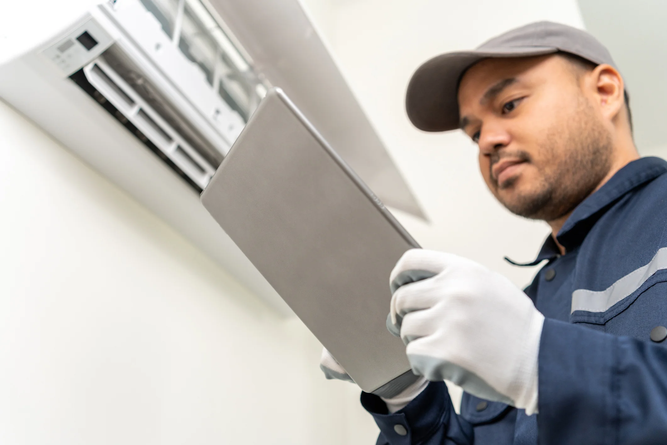 Low-angle view of a technician in gloves inspecting an indoor AC unit and using a digital tablet.