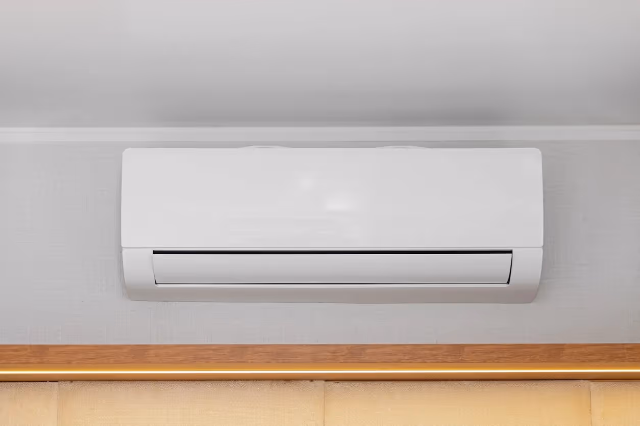 A front-facing view of a sleek, white ductless mini-split air conditioning unit mounted on a light gray wall. The unit is positioned horizontally above a wooden trim and a neutral-toned headboard.