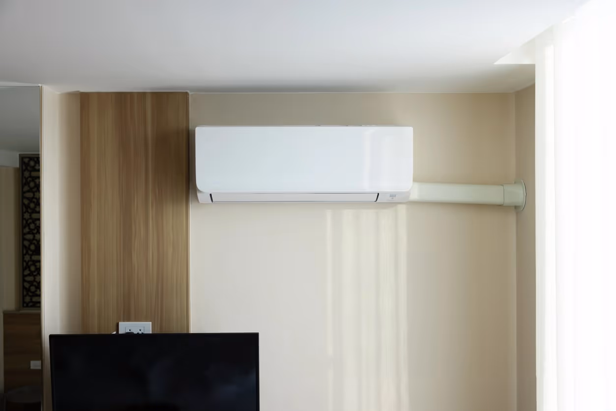 Wall-mounted white AC unit installed above wooden panel and TV in a bright room with sheer curtains.