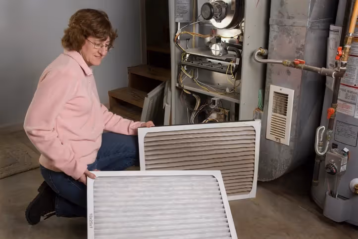 A woman crouches near a furnace, holding a dirty, used air filter next to a clean, new air filter, suggesting maintenance or installation of a new filter.