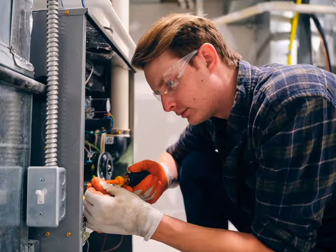 Technician wearing safety glasses and gloves uses a screwdriver to work on the internal electrical components of a furnace, suggesting a repair service.