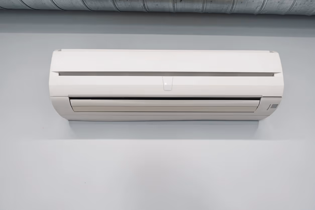 Wall-mounted AC unit on beige wall with exposed ductwork above and a TV partially visible below.